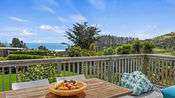 A wooden deck with a table and a bowl of fruit, overlooking a coastal landscape with trees and a view of the sea.