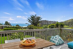 A wooden deck with a table and a bowl of fruit, overlooking a coastal landscape with trees and a view of the sea.