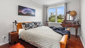A bedroom with a bed, a nightstand, a desk, a chair, a window with curtains, and a wall-mounted mirror.