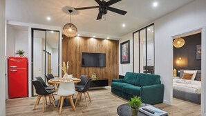 A modern living room with a green sofa, a dining area with a wooden table and chairs, a red refrigerator, and a ceiling fan.