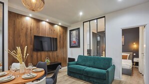A modern living room with a green sofa, dining table set for four, and a flat-screen TV mounted on the wall.