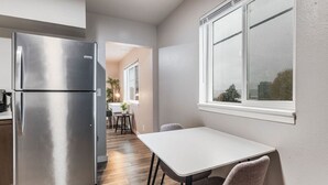A compact kitchen with a stainless steel refrigerator, a white dining table, and a window with a view of the city.