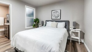 A modern bedroom with a large bed, a bedside table, a nightstand, a window, and a wall-mounted artwork.