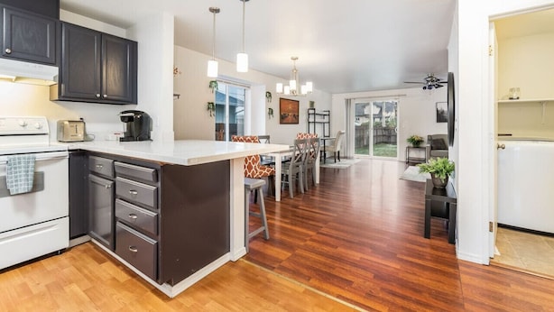 Una cocina moderna con armarios oscuros, una encimera blanca y suelo de madera. Hay una zona de comedor con una mesa y sillas.