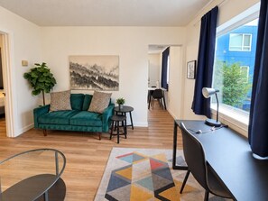 A living room with a green sofa, a small round table, a desk with a chair, and a window with curtains.