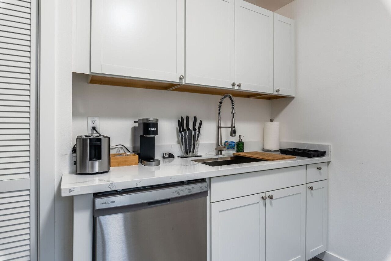 A compact kitchen with white cabinets, a built-in dishwasher, a coffee maker, and a sink with a faucet.
