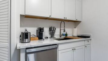 A compact kitchen with white cabinets, a built-in dishwasher, a coffee maker, and a sink with a faucet.