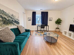 Un salon avec un canapé vert, une table basse en verre, un bureau avec une chaise, une plante en pot et un téléviseur posé sur un meuble blanc.