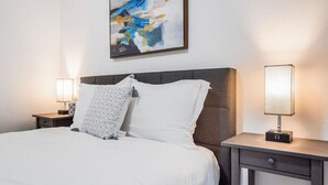 A neatly made bed with white linens and two pillows, a modern abstract painting on the wall, and two bedside lamps on nightstands.