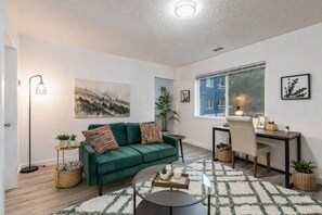 Un salon moderne avec un canapé vert, une table basse ronde, un bureau et une grande fenêtre.