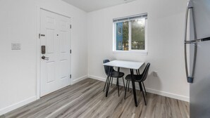 A small, bright kitchen with a white door, a window, a table with two chairs, and a refrigerator.