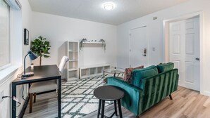 A modern living room with a green sofa, a small round table, a desk with a lamp, and a white shelving unit.