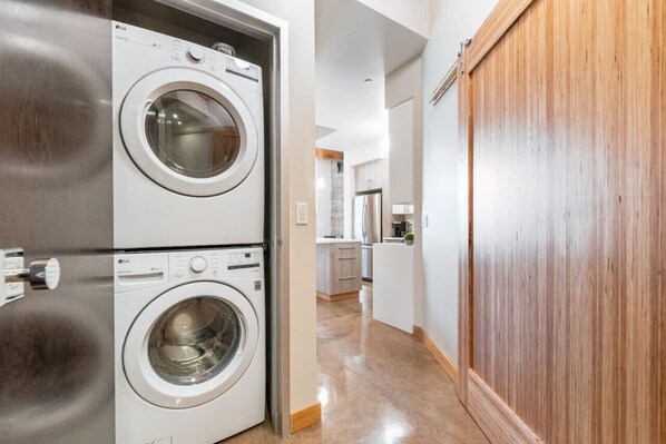 A laundry room with two stacked washing machines, a wooden sliding door, and a kitchen area in the background.
