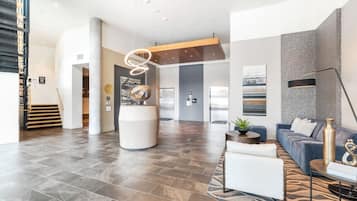 A spacious lobby with a modern design, featuring a central seating area with sofas and a coffee table, a staircase on the left, and a large pillar with a unique light fixture.