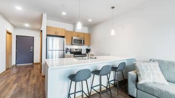 Una cocina moderna con una isla central, taburetes de bar y un sofá gris en una sala de estar luminosa.