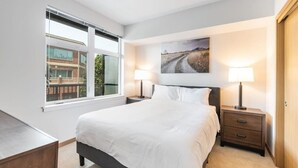 Ein Schlafzimmer mit einem großen Bett, zwei Nachttischen mit Lampen, einem Fenster mit Blick auf ein Gebäude und einem Gemälde an der Wand.