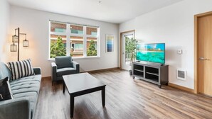 Ein modernes Wohnzimmer mit großem Flachbildfernseher, einer Couch, einem Couchtisch und einem Balkon mit Blick auf ein Gebäude und Grünflächen.