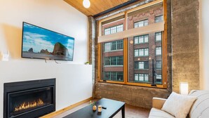 A modern living room with a fireplace, a television mounted on the wall, and a large window overlooking a brick building.