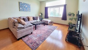 A living room with a sectional sofa, a coffee table, a ceiling fan, a large window with curtains, and a television on a stand.