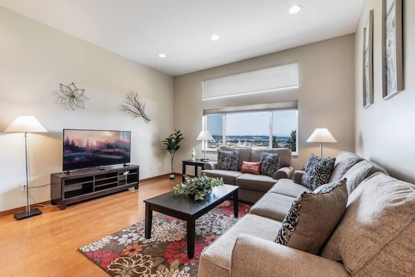 A modern living room with a flat-screen TV, a sofa set, a coffee table, and a large window offering a view.