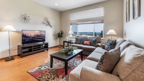 A modern living room with a flat-screen TV, a sofa set, a coffee table, and a large window offering a city view.