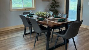 Une salle à manger avec une table en bois dressée pour six personnes, entourée de six chaises. Il y a deux fenêtres donnant sur les montagnes, un tableau encadré au mur et un petit arbre dans un coin.