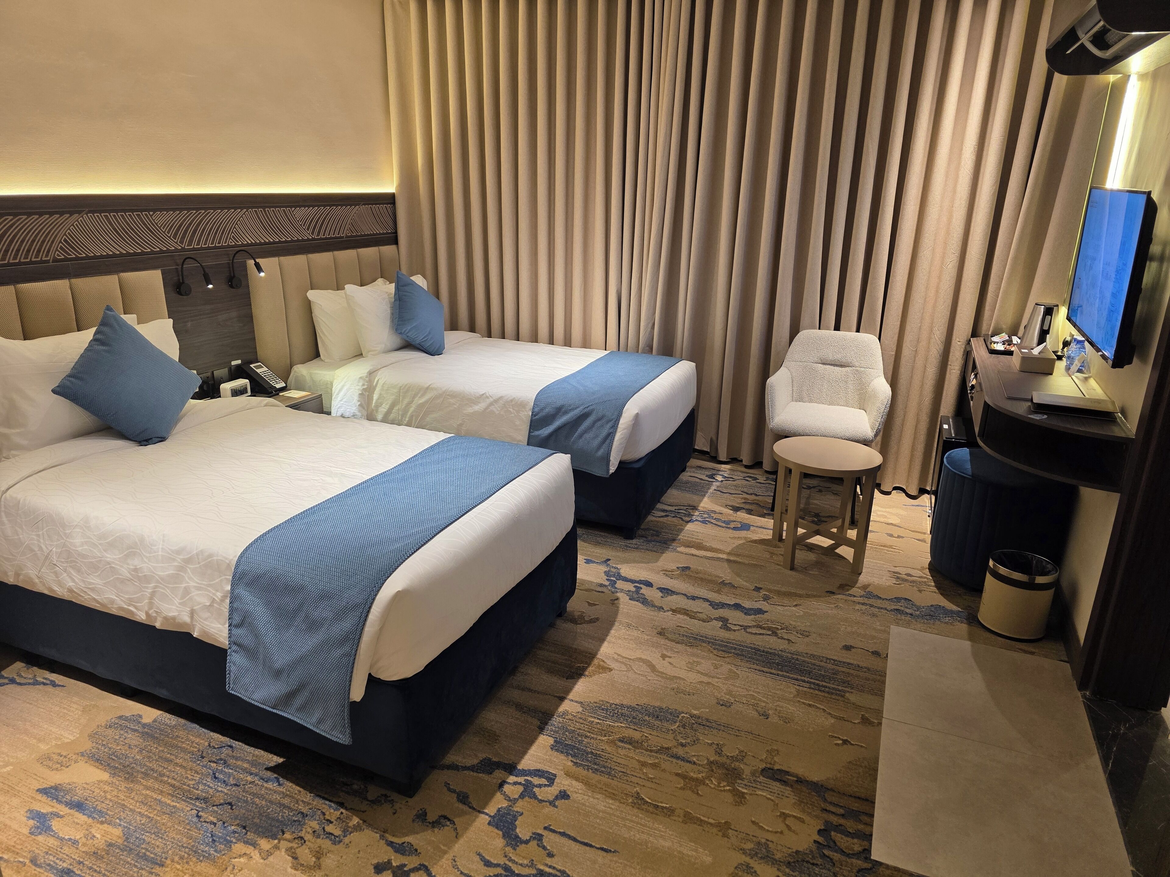 A hotel room with two beds, a desk, a chair, a TV, and a trash can.
