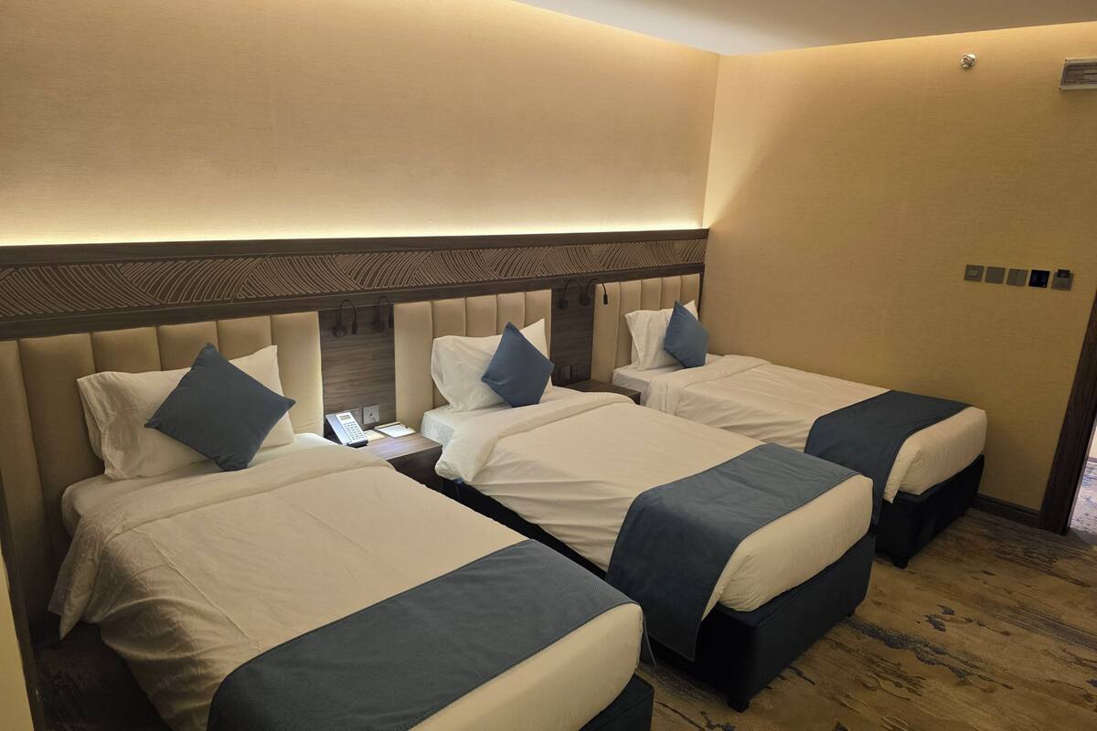 Habitación de hotel con tres camas dispuestas en fila, cada una con cabecero y almohadas. Entre las camas, un mesita de noche con un teléfono y una lámpara.