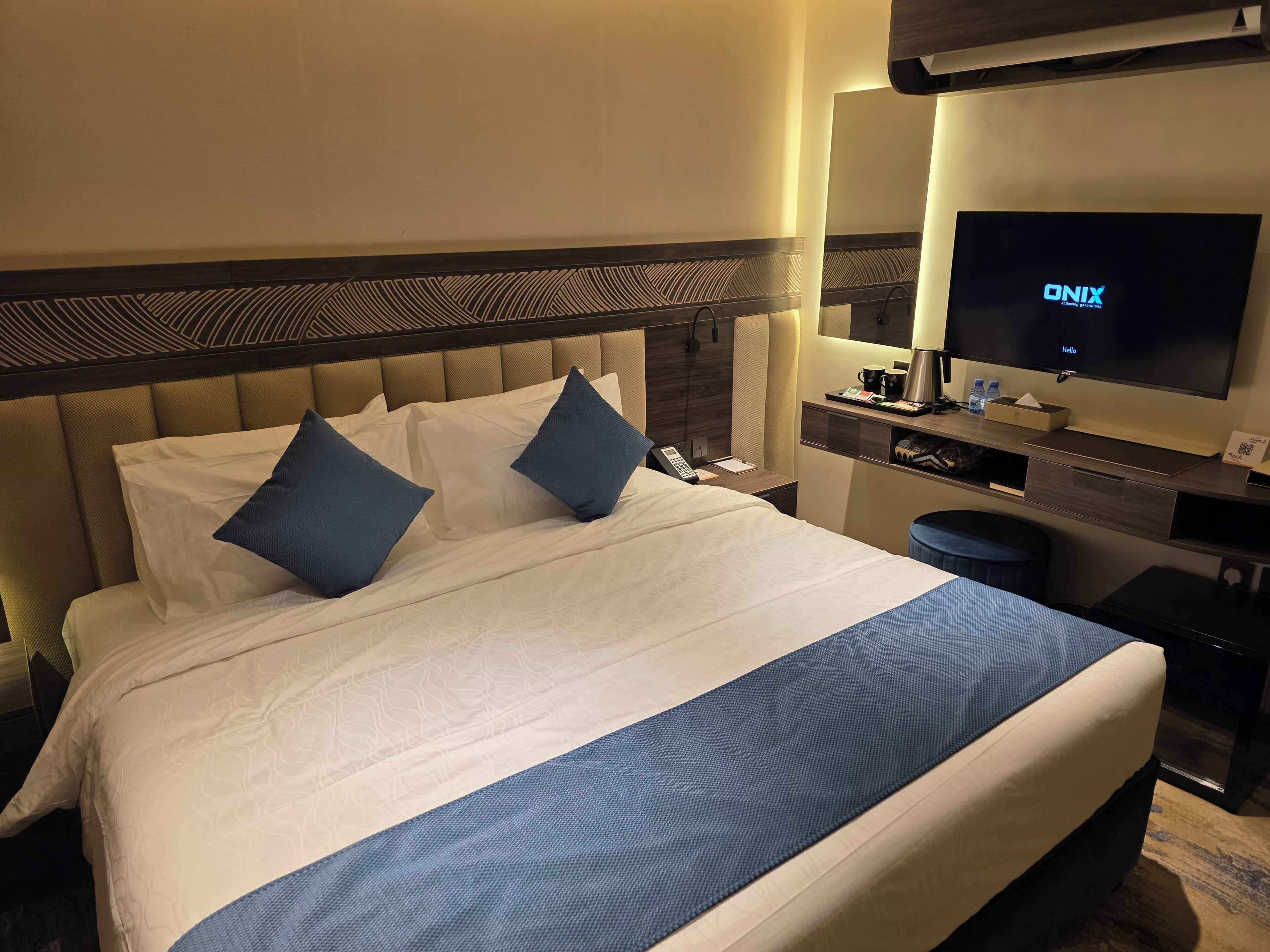 A hotel room with a large bed, a television, a bedside table with a phone, and a small desk area with a coffee maker and a tray.