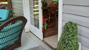 Varanda coberta com móveis de vime, um vaso de planta e uma área de refeições visível pela porta de vidro.