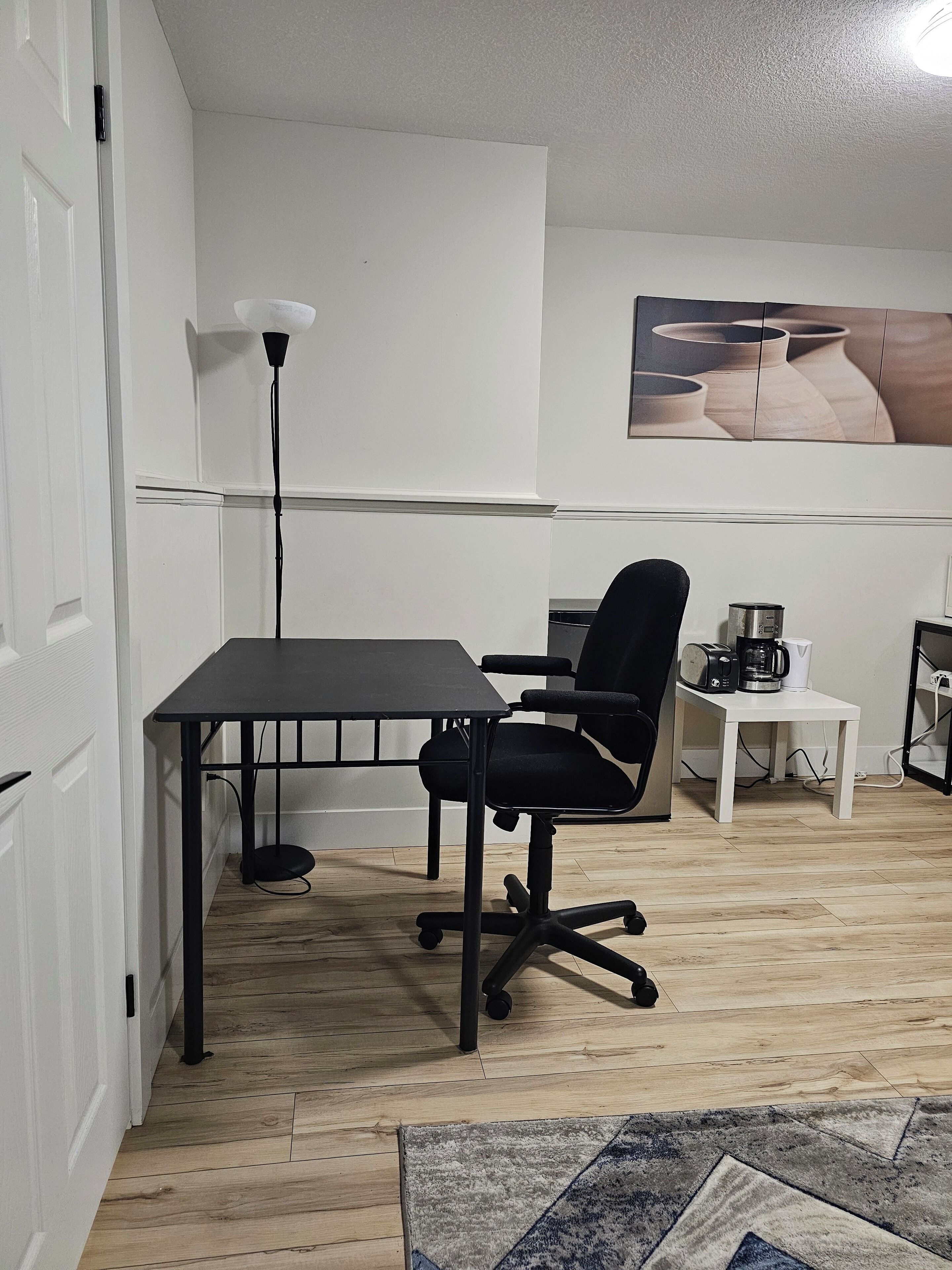 A small, modern room with a wooden floor, a black office chair, a white table, and a coffee maker.