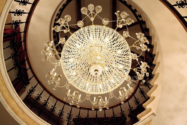 A grand, ornate chandelier with a circular, floral design, suspended in a spacious, elegantly decorated room with a spiral staircase.