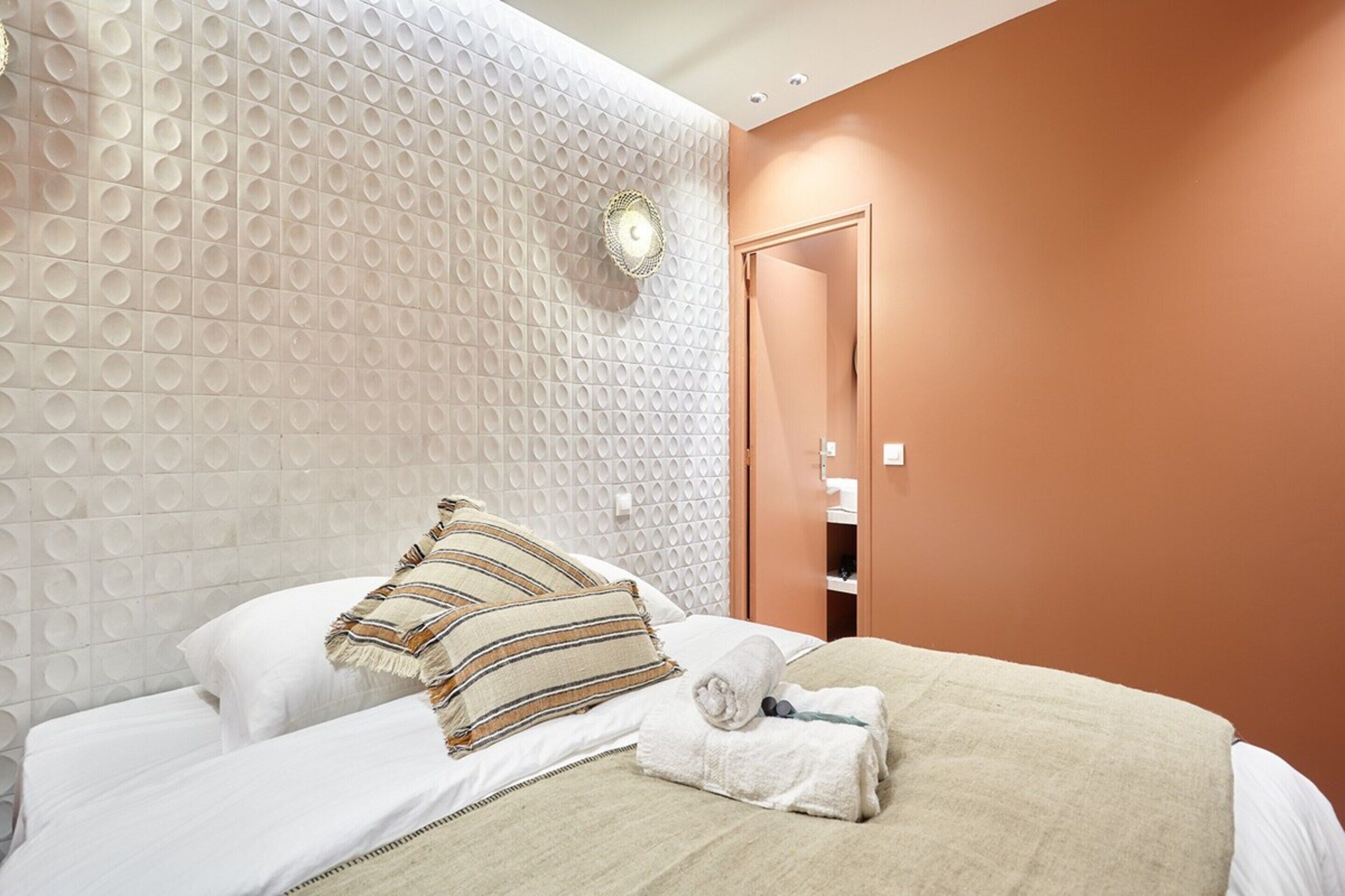 A neatly made bed with white linens and striped pillows, a beige blanket, and a rolled towel.