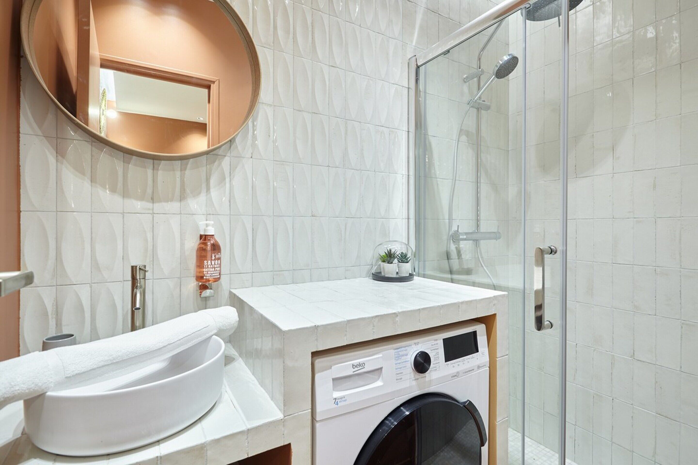 A modern bathroom with a round mirror, a sink, a washing machine, and a glass-enclosed shower.