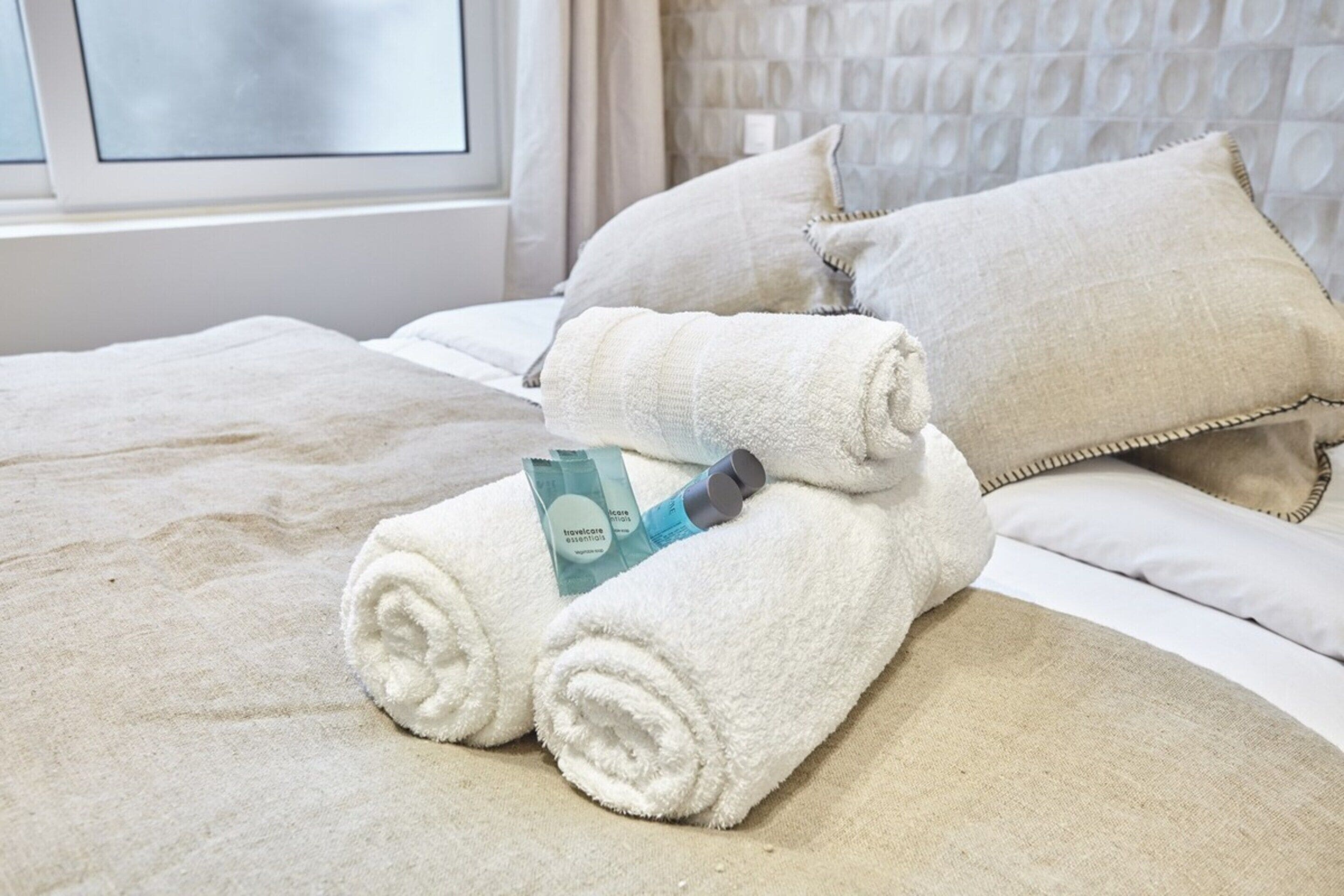 A neatly made bed with white linens, a rolled towel, and a travelcare product on the bed.