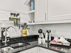 Una cocina con un lavabo, una cafetera y un hervidor. Hay utensilios colgados en la pared y una balanza en la encimera.