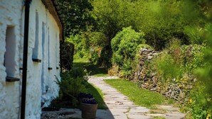 Un sendero de piedra conduce a un edificio blanco con techo inclinado, rodeado de abundante vegetación y un muro de piedra.