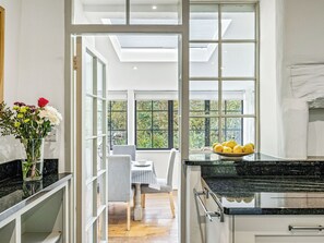 Una cocina con encimera negra, un comedor con mesa y sillas, y vistas a zonas verdes a través de amplios ventanales.