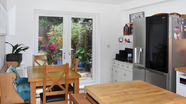 A modern kitchen with a wooden dining table, chairs, a refrigerator, and a window overlooking a garden.