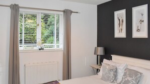 A bedroom with a bed, a window with curtains, a radiator, a bedside table with a lamp, and framed botanical prints on the wall.