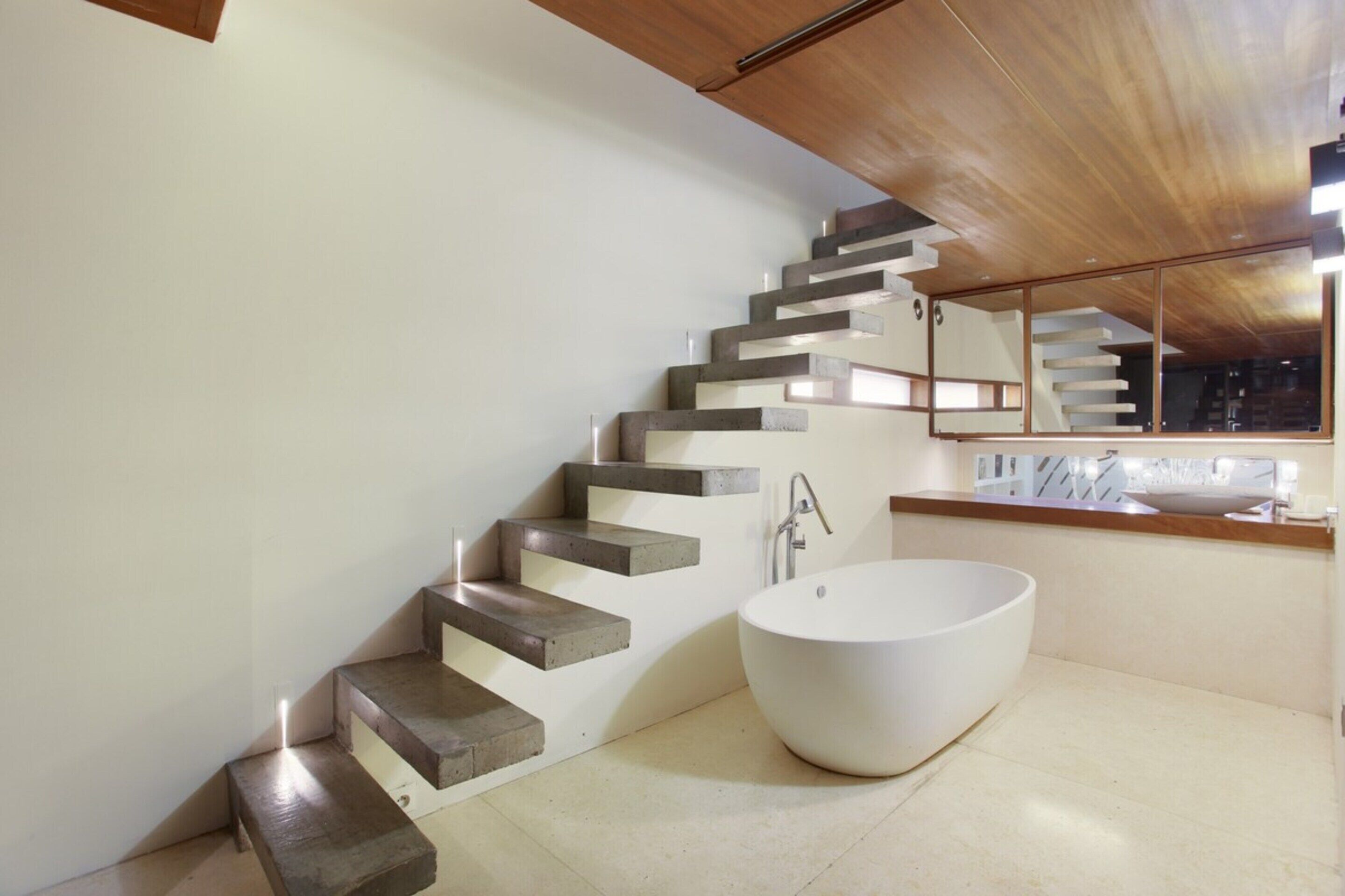 A modern bathroom with a freestanding bathtub, a wooden staircase, and a large mirror.