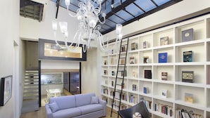 Una sala de estar moderna con un sofá gris, una mesa de centro de madera y una silla negra. Hay una gran estantería blanca llena de libros y objetos enmarcados. Un candelabro cuelga del techo, y una escalera está apoyada contra la estantería.