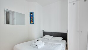 Una cama bien hecha con sábanas blancas y una pila de toallas dobladas. Un espejo grande está montado en la pared sobre la cama.