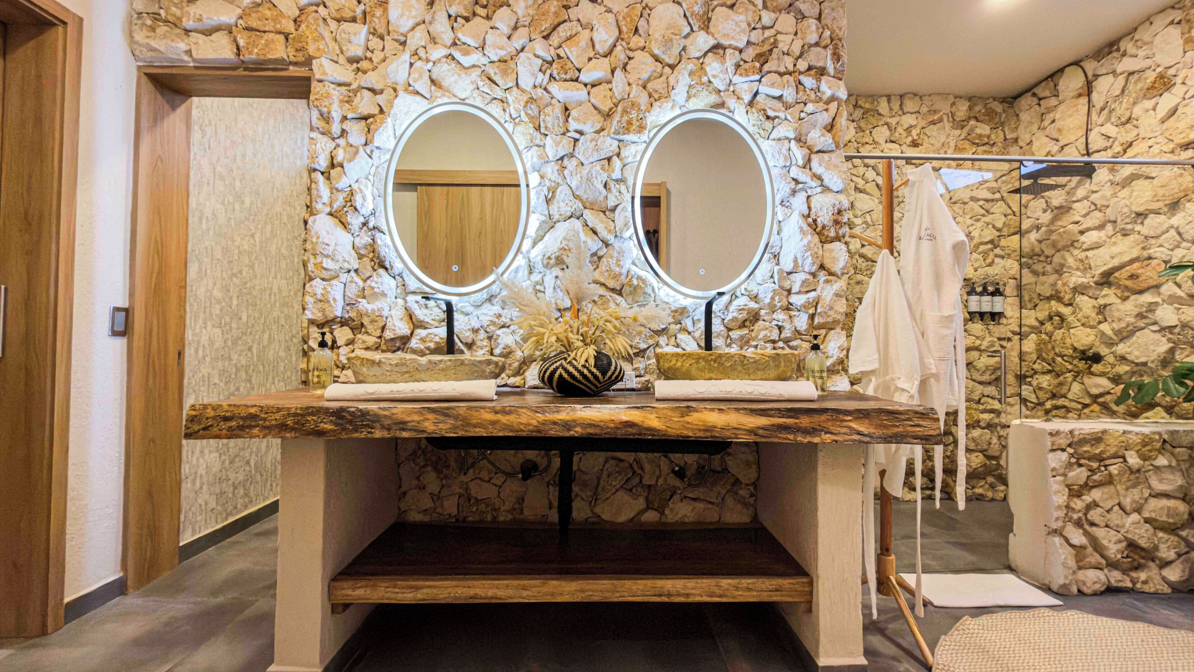 A bathroom with a stone wall, two mirrors, a sink with a vase, and a shower area.