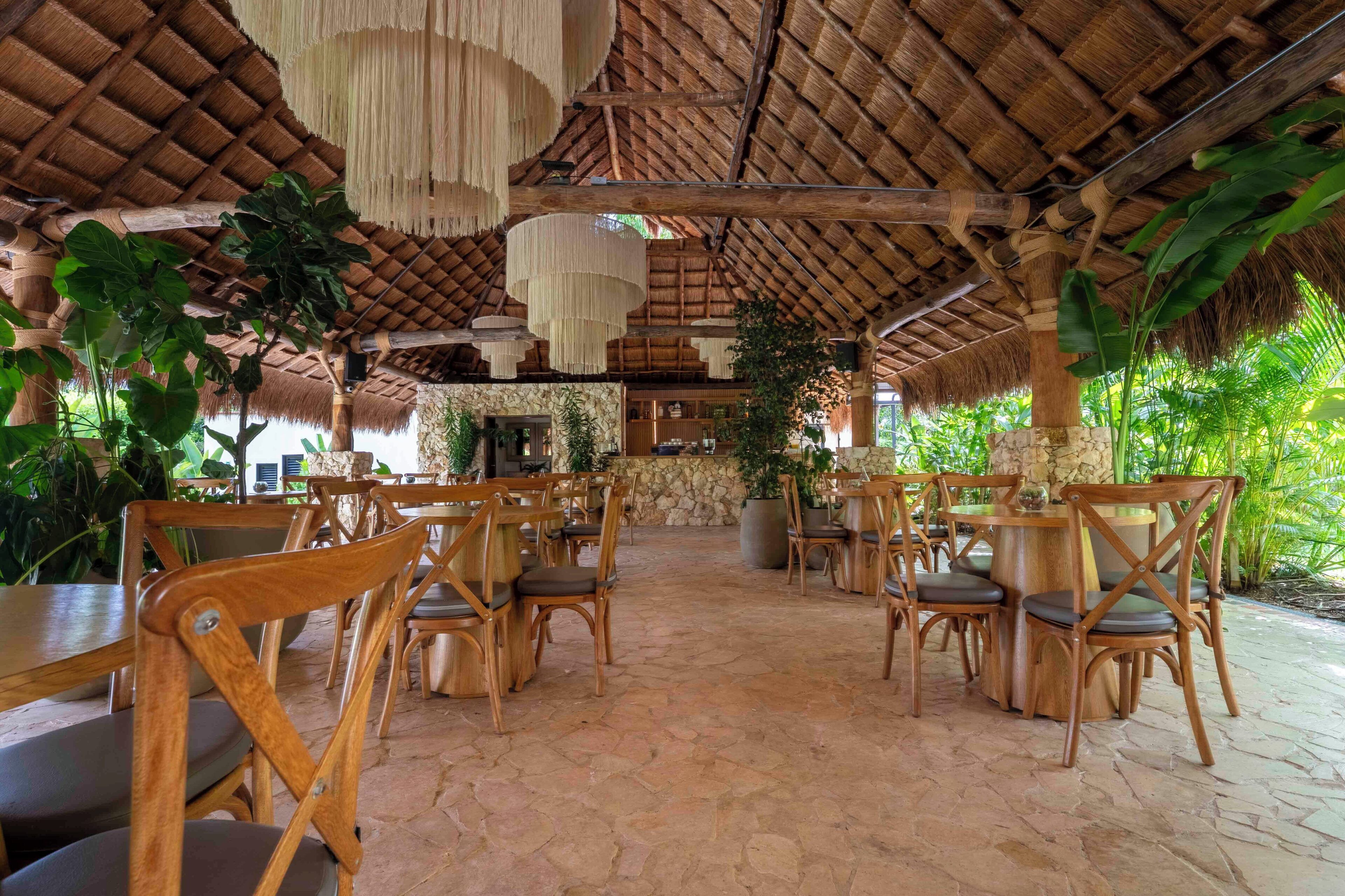A covered outdoor dining area with wooden chairs and tables, a stone floor, and hanging light fixtures.