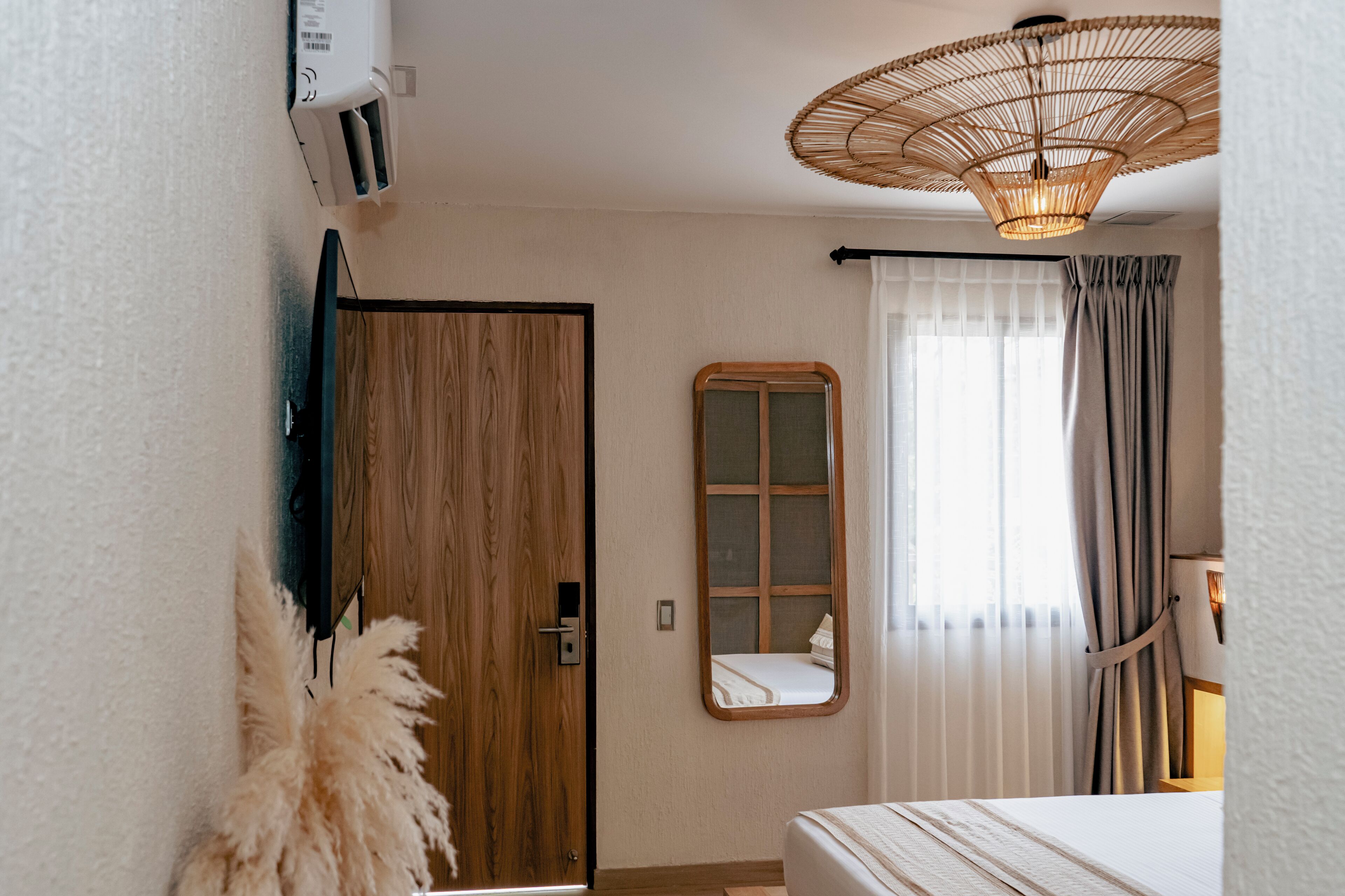 Habitación de hotel con puerta de madera, espejo, cama, ventilador de techo y ventana con cortinas.
