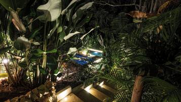A nighttime garden with a pool, surrounded by lush tropical plants and illuminated by soft lighting.
