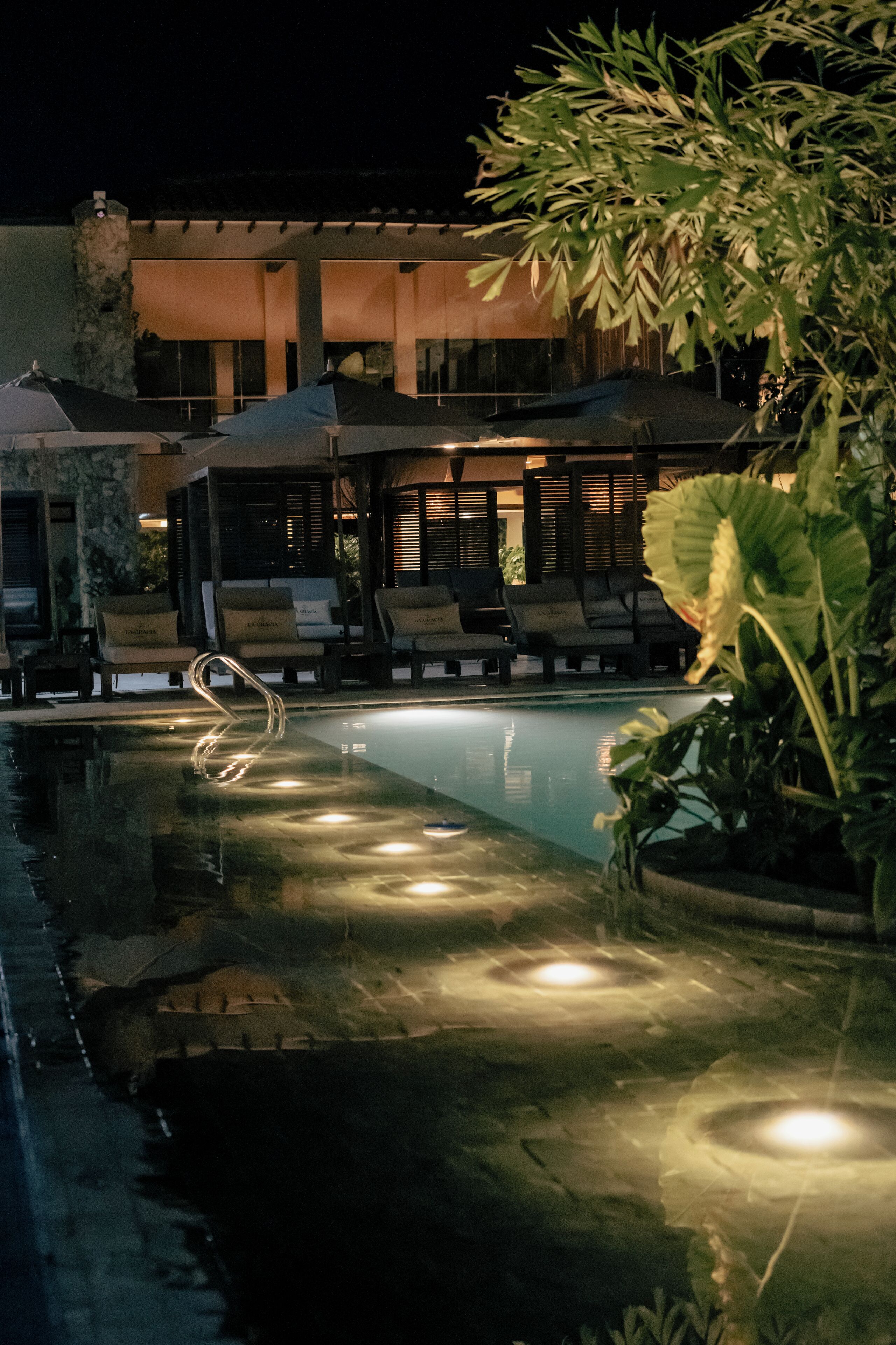 A nighttime pool area with lounge chairs and umbrellas, surrounded by greenery.