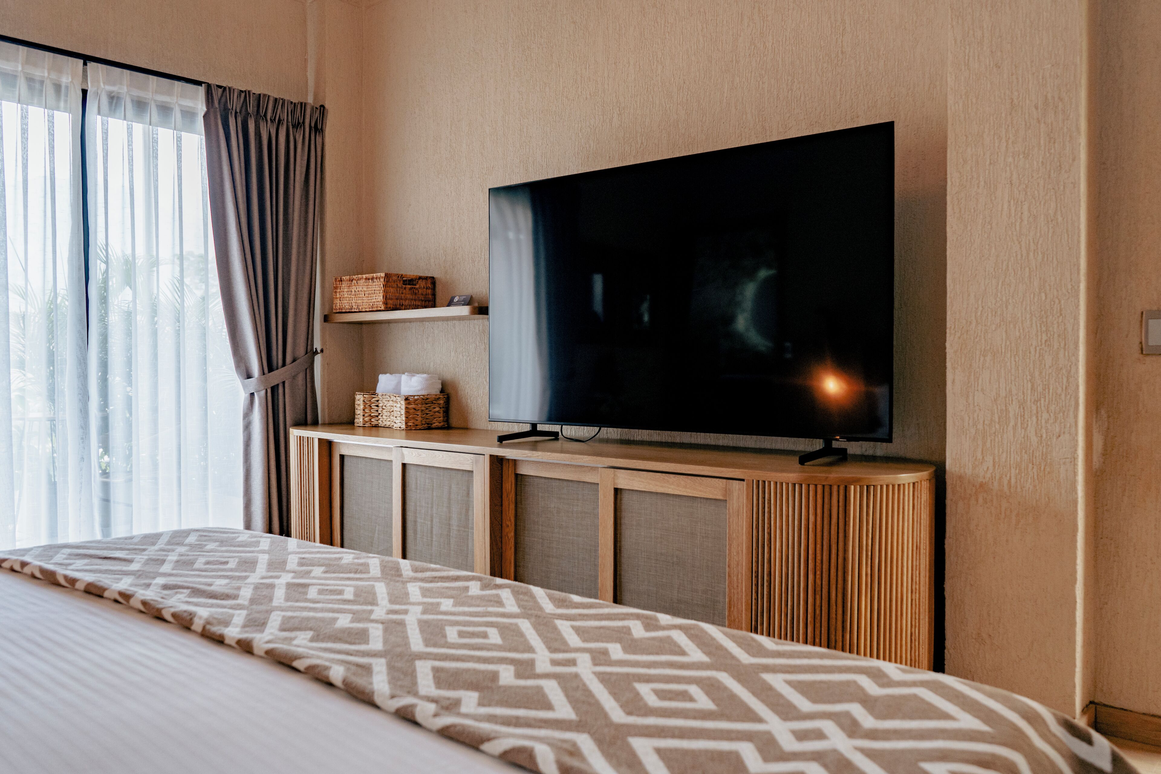 Habitación de hotel con una cama grande, un televisor de pantalla plana montado en un mueble de madera, una ventana con cortinas y un pequeño estante con adornos.