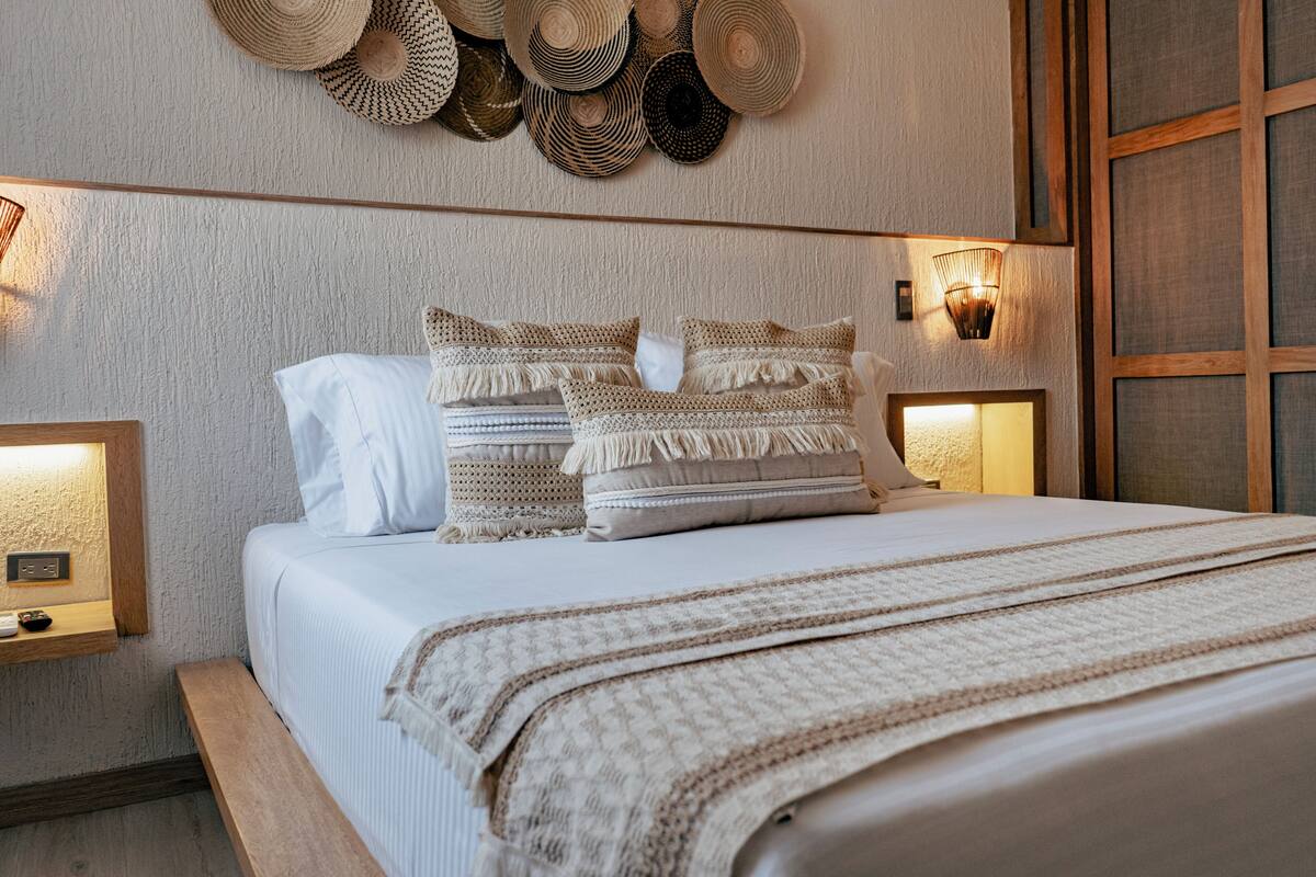 Una cama bien hecha con una colcha blanca y un edredón color beige, flanqueada por dos mesitas de noche con lámparas.